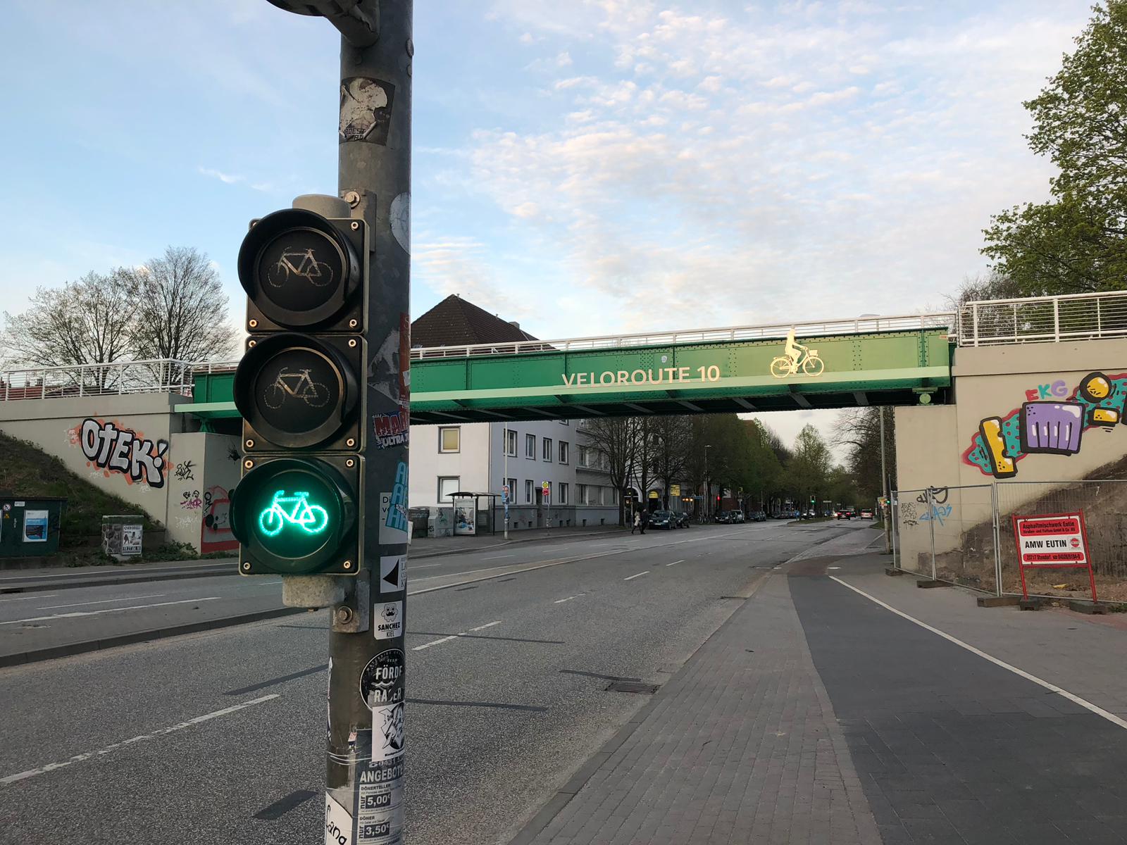 grüne Brücke über einer Straße mit der Aufschrift Veloroute 10, im Vordergrund eine Ampel, die auf grünem Fahrrad steht