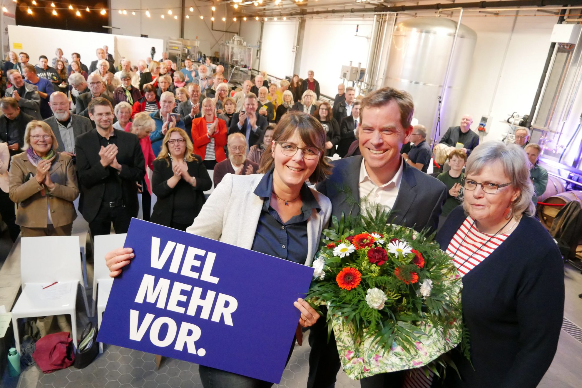 Gesine Stück (mit einem Schild "viel mehr vor"), Ulf Kämpfer (mit Blumenstrauß) und Gesa Langfeldt auf der Bühne. Hinter ihnen stehen die Gäste und klatachen.