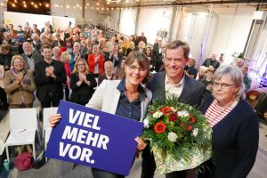 Gesine Stück (mit einem Schild "viel mehr vor"), Ulf Kämpfer (mit Blumenstrauß) und Gesa Langfeldt auf der Bühne. Hinter ihnen stehen die Gäste und klatachen.