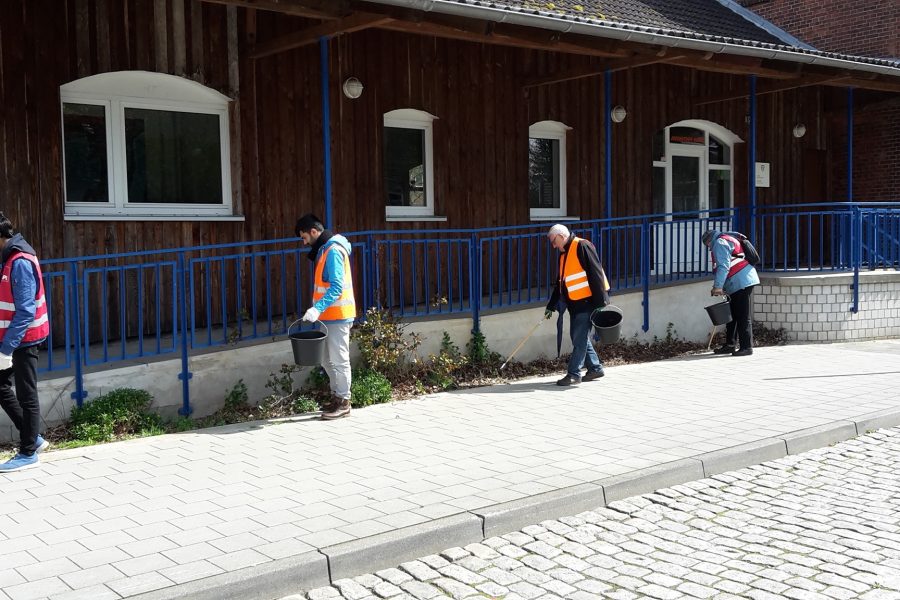 4 Männer mit orangen Warnwesten sammeln Müll an einer Rampe vor einem Haus mit Holzfassade