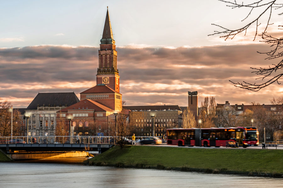 Dieses Foto zeigt den kleinen Kiel im Vordergrund und den Kieler Rathausturm im Hintergrund bei Abenddämmerung sowie einen fahrenden Bus leicht unscharf in Bewegung...