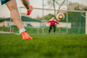 Beine eines Mannes, der einen Fußball gerade ins Tor schießt. Im Hintergrund unscharf der Torwart im Tor auf einem Raseplatz.