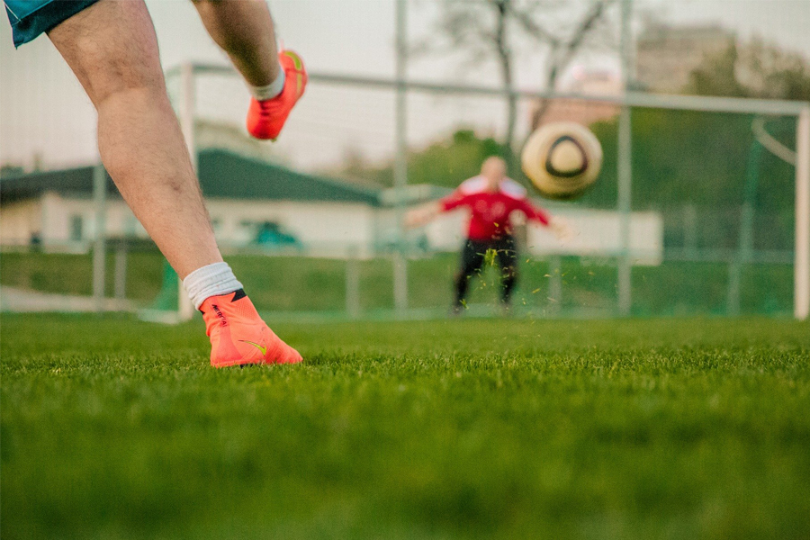Foto zeigt einen Mann, der den Fußball gerade ins Tor schießt und im Hintergrund unscharf den Torwart im Tor auf dem Fußballplatz im Freien.