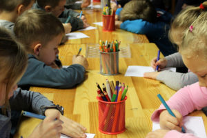 Mehrere Kinder sitzen an einem langen Tisch und malen mit Bundstiften auf Papier