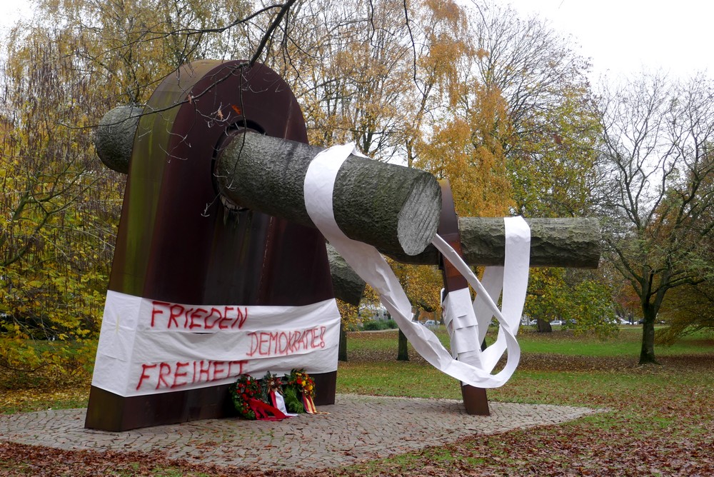 Breuste-Denkmal mit Papierbahnen umwickelt auf denen steht: Frieden, Demokratie, Freiheit. Davor liegen drei Kränze.
