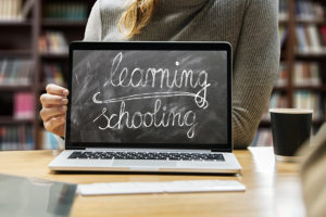 Display eines Laptop auf dem mit Kreide geschrieben steht "learning schooling". Im Hintergrund Frau, die den Laptop hält und ein Bücherregal