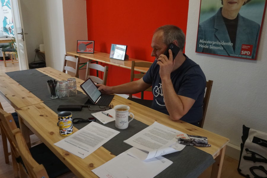 Mathias stein telefoniert im Bürgerbüro, guckt gleichzeitig auf sein Tablet. Hinter ihm ist ein Plakat von Heide Simonis, neben ihm eine Tasse Kaffee und mehrere Unterlagen.