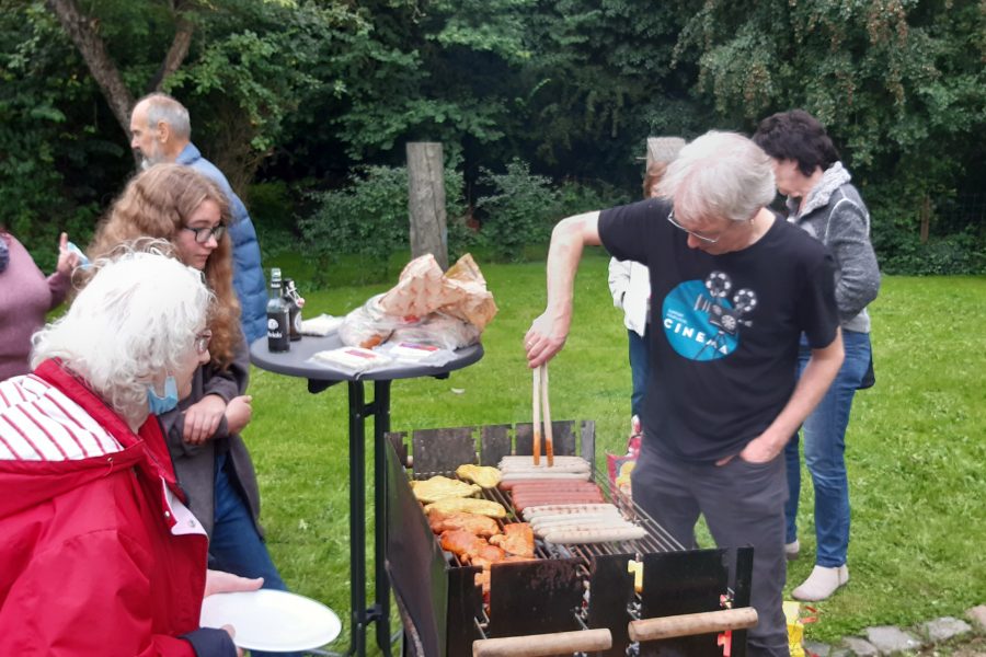 Ein Mann steht mit einer Zange an einem Grill mit Würstchen. Daneben steht eine Frau mit einem Teller in der Hand