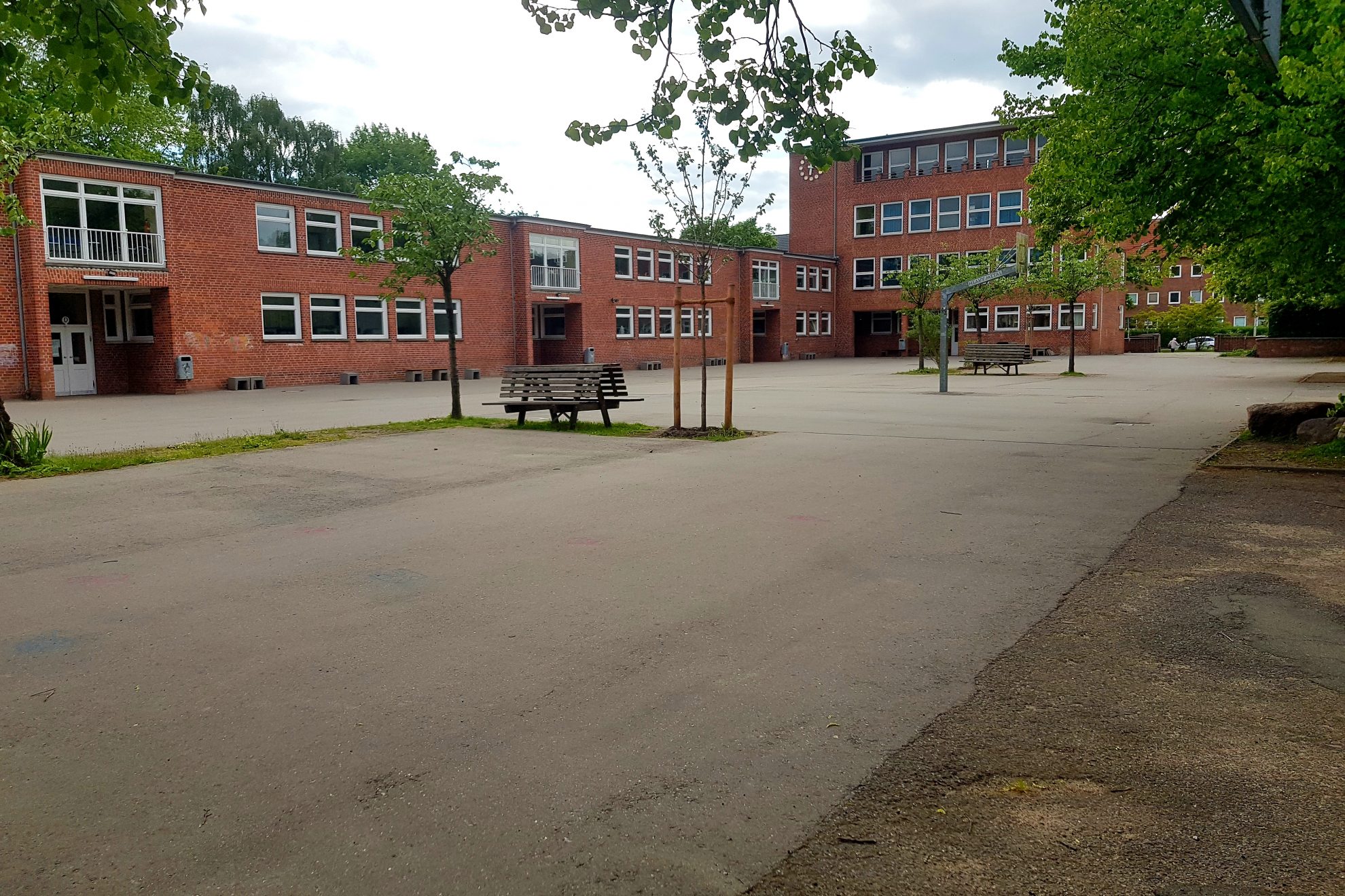 Schulhof mit Schulgebäude (roter Backsteinbau). Auf dem Schulhof sind zwei Bänke und ein paar kleine Bäume.