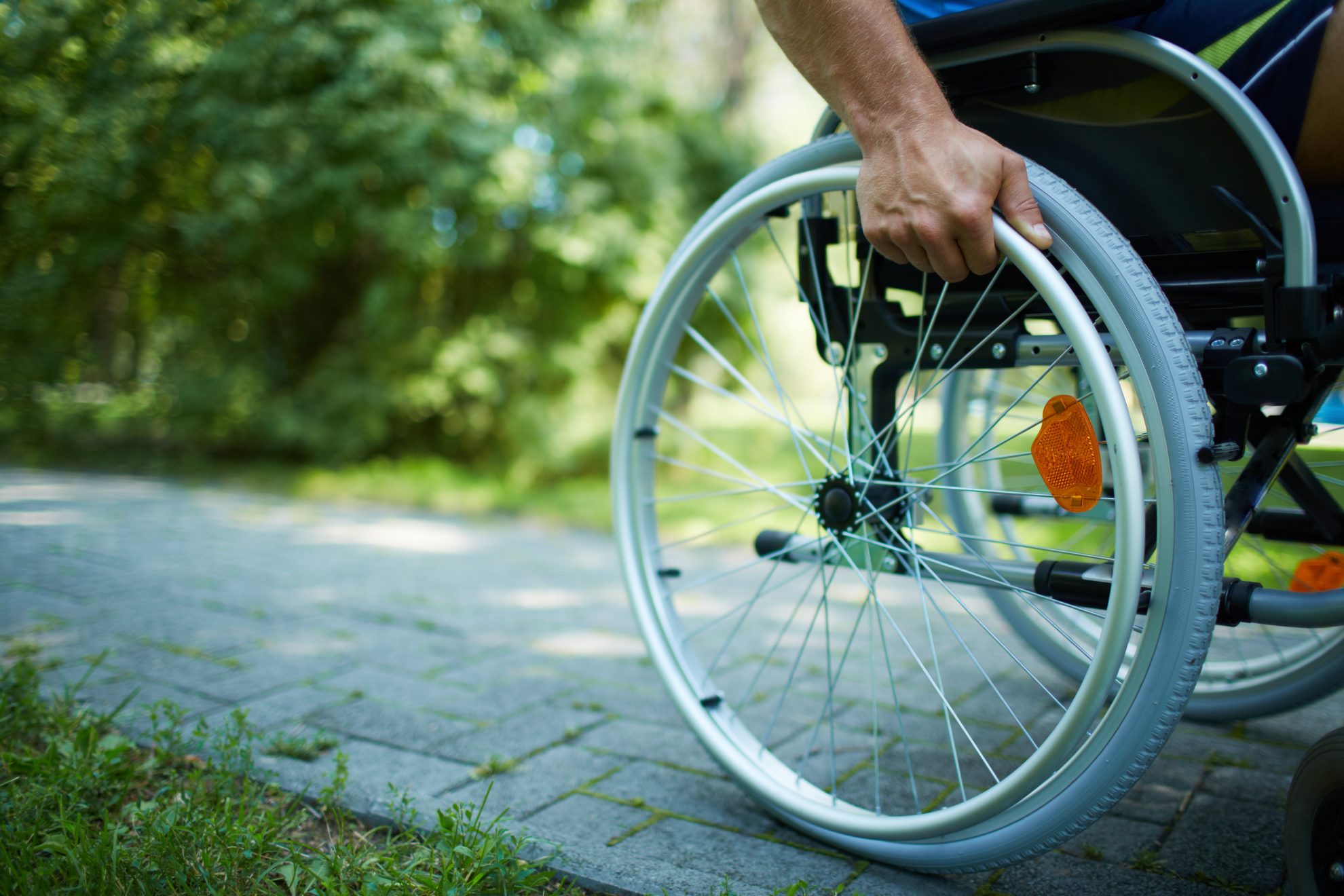 Mann fährt auf einem Rollstuhl auf einem Gehweg. Zu sehen ist vor allem ein Rad und eine Hand. Im Hintergrund zu sehen ist Grün.