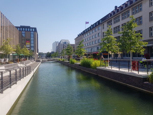 Kanal auf beiden Seiten Bäume und Gebäude in der Ferne eine Brücke