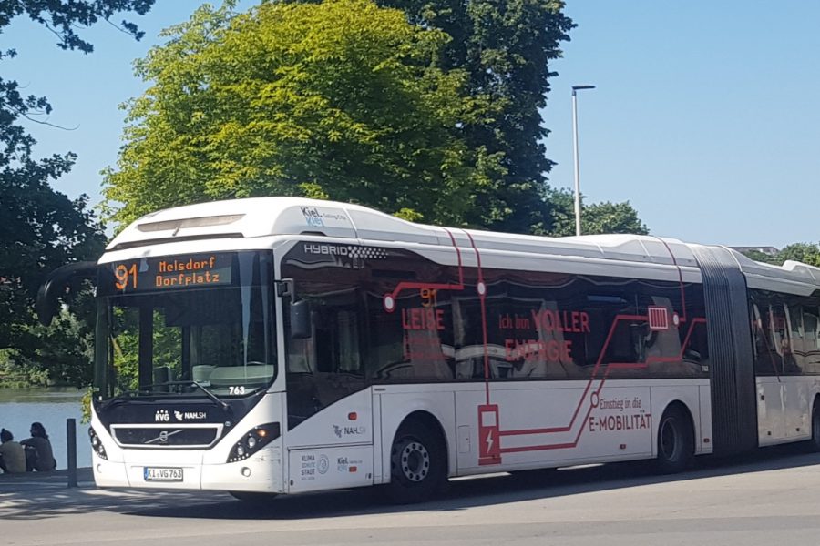 Weißer Elektrobus (Anzeige 91 Melsdorf Dorfplatz) auf einer Straße, im Hintergrund Bäume und Himmel