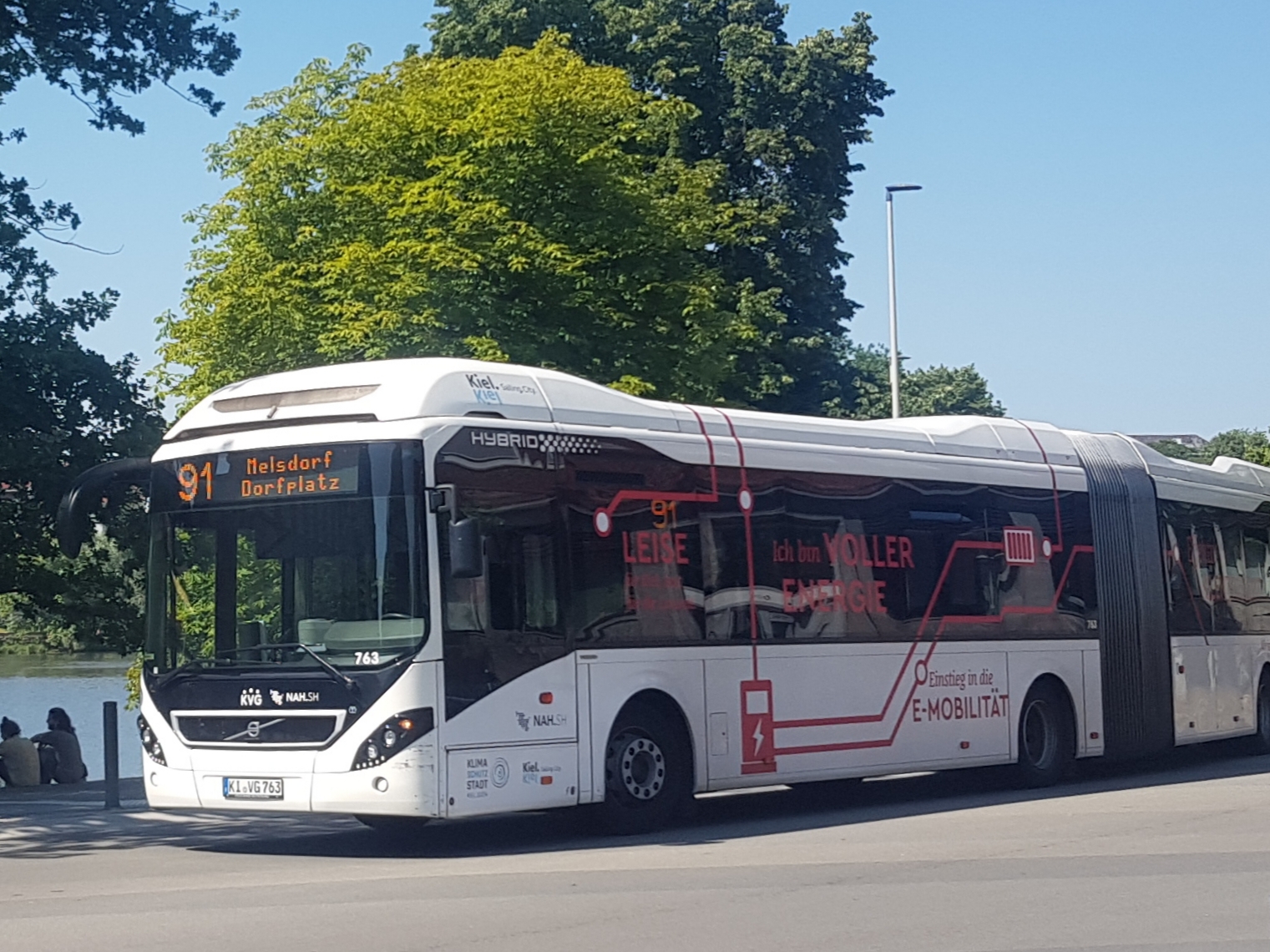 Weißer Elektrobus (Anzeige 91 Melsdorf Dorfplatz) auf einer Straße, im Hintergrund Bäume und Himmel