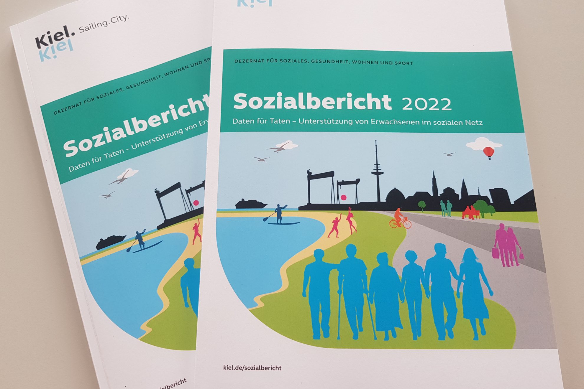 Zwei Broschüren, auf denen vorne steht "Sozialbericht 2022: Daten für Taten: Unterstützung von Erwachsenen im sozialen Netz". Links oben steht "Kiel. Sailing City".