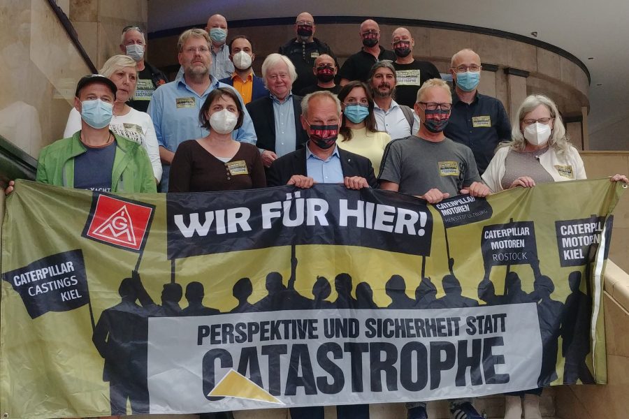 Die SPD-Ratsfraktion steht gemeinsam mit den Betriebsräten von Caterpillar auf der Treppe im Kieler Rathaus und hält ein Solidaritätsbanner