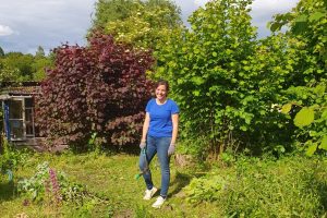Christina Schubert steht im Garten, hat Gartenhandschuhe an und eine Hacke in der Hand.