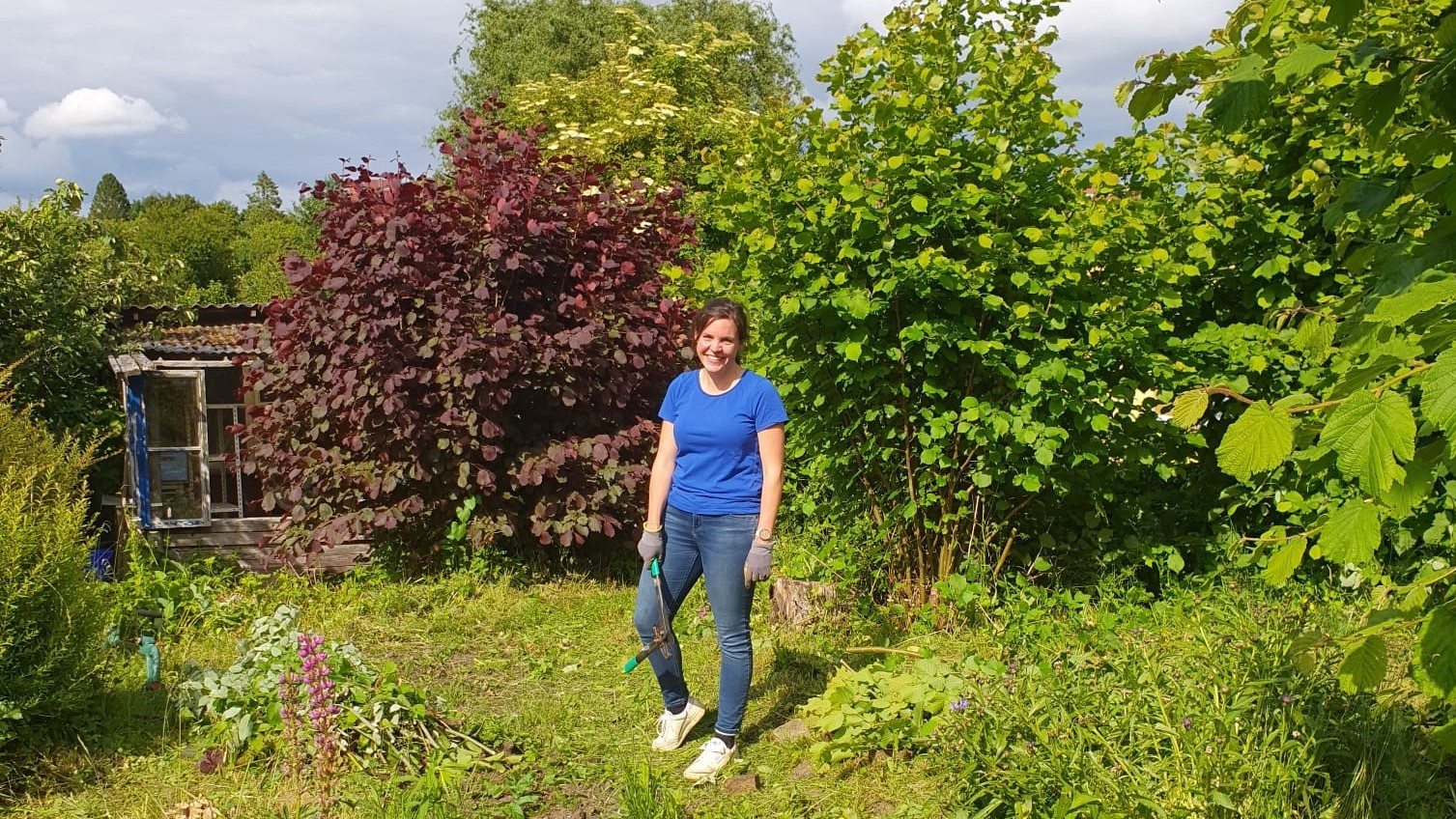 Christina Schubert steht im Garten, hat Gartenhandschuhe an und eine Hacke in der Hand.