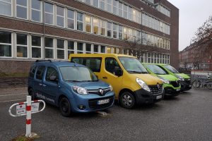 Vier große Autos, darunter ein Transporter stehen nebeneinander. Auf dem einen ist der Schriftzug "StattAuto Carsharing" erkennbar. Im Hintergrund ist das Arbeitsamtsgebäude am Wilhelmplatz zu sehen.