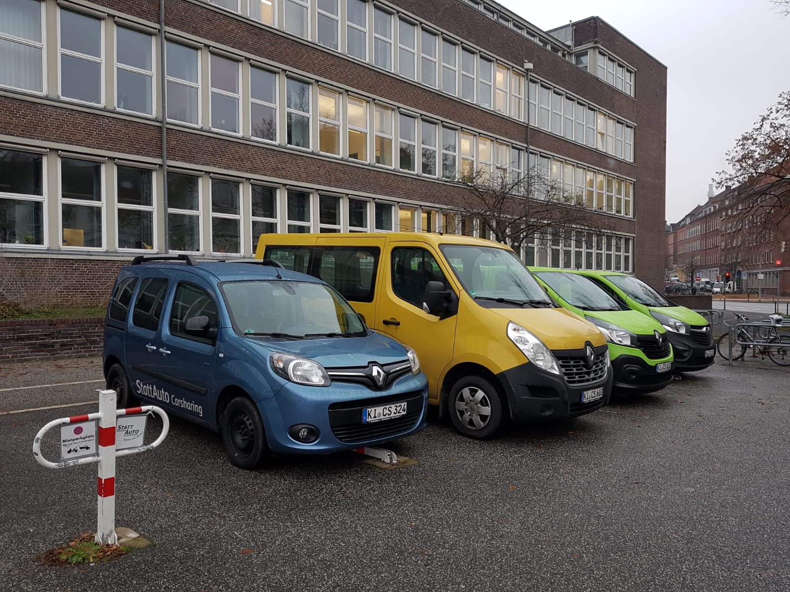 Vier große Autos, darunter ein Transporter stehen nebeneinander. Auf dem einen ist der Schriftzug "StattAuto Carsharing" erkennbar. Im Hintergrund ist das Arbeitsamtsgebäude am Wilhelmplatz zu sehen.