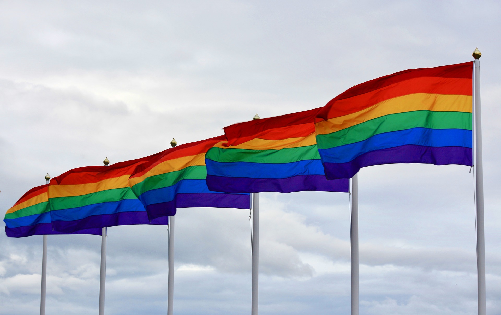 Fünf Regenbogenflaggen wehen nebeneinander, im Hintergrund wolkiger Himmel.