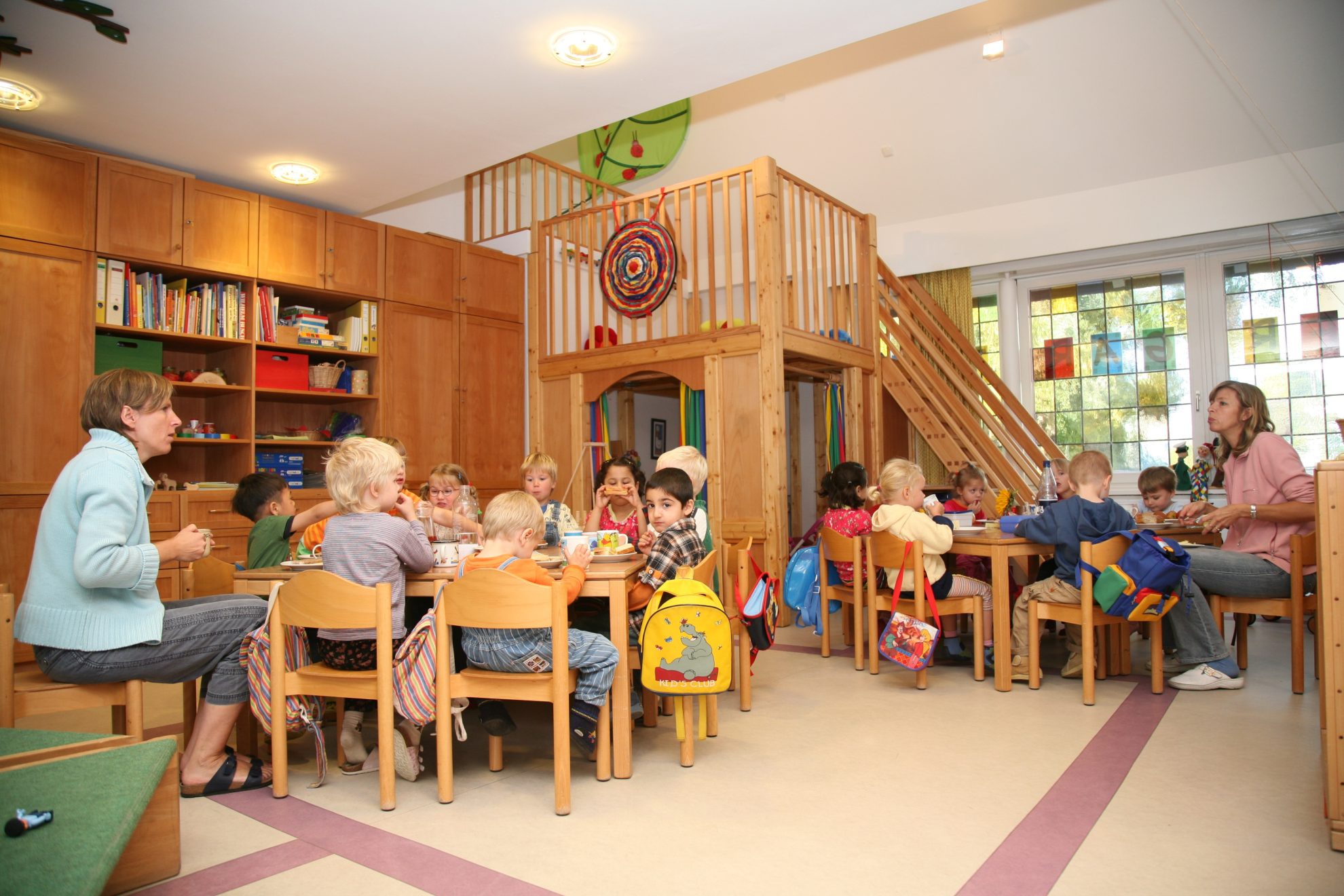 Eine Gruppe Kinder sitzt auf keinen Stühlen an kleinen Tischen in einer Kita. Rechts und links sitzt jeweils eine Erzieherin.