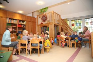 Eine Gruppe Kinder sitzt auf keinen Stühlen an kleinen Tischen in einer Kita. Rechts und links sitzt jeweils eine Erzieherin.