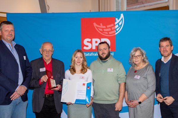 Sechs Menschen stehen vor einer blauen Rückwand mit dem Logo der SPD-Ratsfraktion. eine Person hält eine Urkunde, die andere einen Pokal