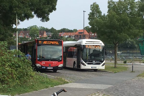 Zwei Busse fahren nebeneinander her. Der linke Bus ist ein alter, roter Bus mit Verbrennungsmotor. Der rechte Bus ist ein neuer weißer Elektrobus.