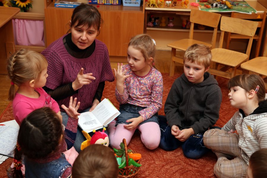 Erzieherin sitzt mit sechs Kindern im Sitzkreis auf dem Boden