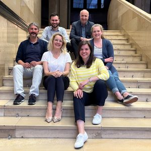 Sechs Personen sitzen auf einer Treppe. Obere Reihe von links nach rechts: Özgürcan Baş, Volhkard Hanns, Mittlere Reihe von links nach rechts: Nesimi Temel, Antje Möller-Neustock, Anna-Lena Walczak, vorne: Christina Schubert