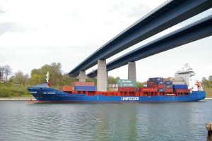 Zwei Brücken führen über eine Wasserstraße. Darunter fährt ein blaues Containerschiff