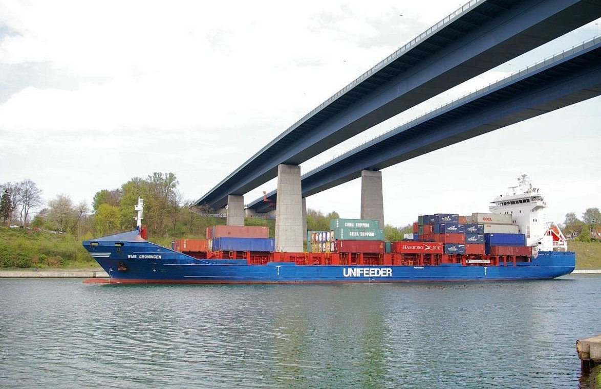 Zwei Brücken führen über eine Wasserstraße. Darunter fährt ein blaues Containerschiff